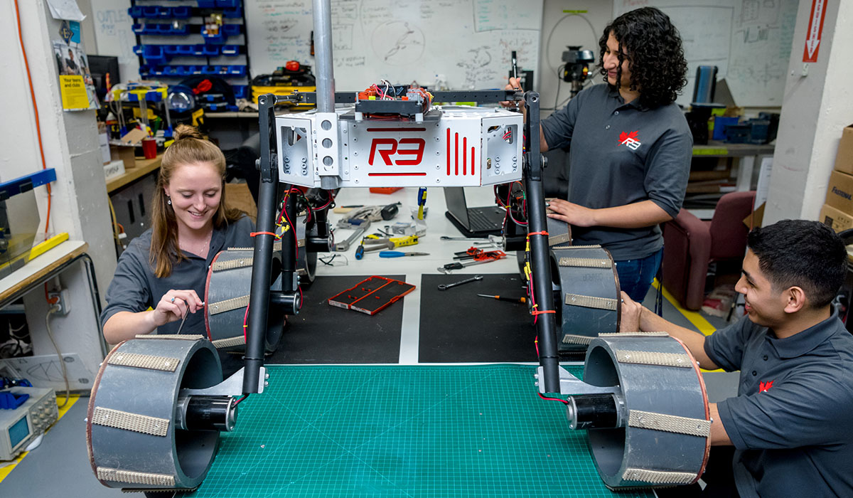 Three Toronto MetRobotics team, TMR, members smiling and working on the next robot for the team in their lab