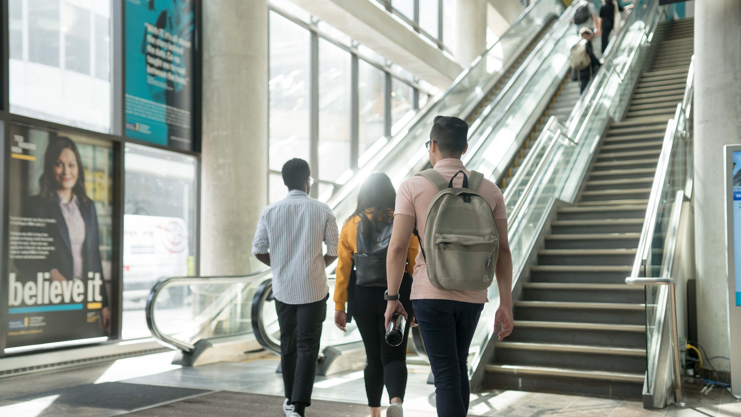 Students walking towards the escalators inside Ted Rogers School of Management building as the sun shines throuogh the windows left of the escalators