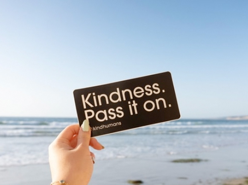 Hand holding a card that reads "Kindess. Pass it on." and the ocean in the background.