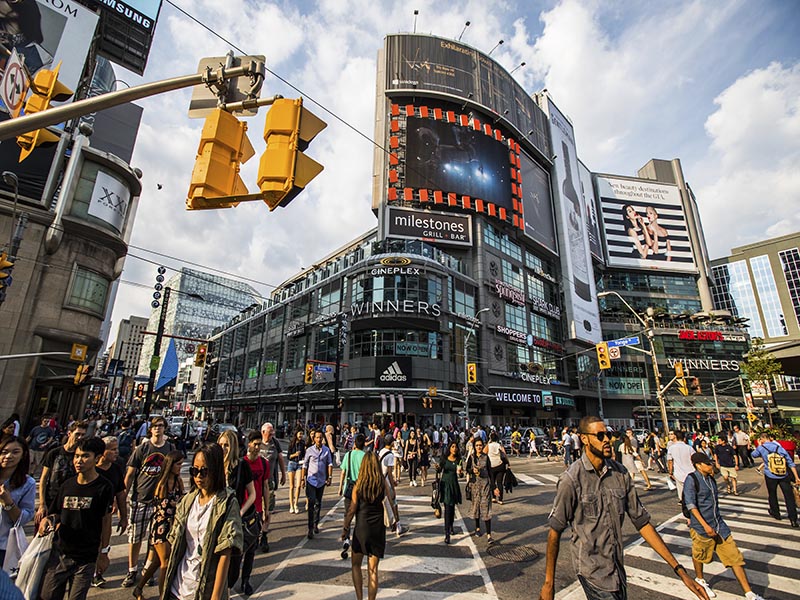 The downtown Toronto intersection Yonge-Dundas Square on a busy summer day. A crowd of people walk across the intersection from all directions with lit up billboards and clouds against a pale blue sky. 