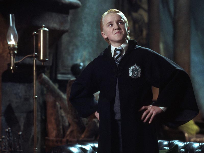 Young Draco Malfoy stands in his black Slytherin robes with his hands on his hips and looking smugly at something to his left.