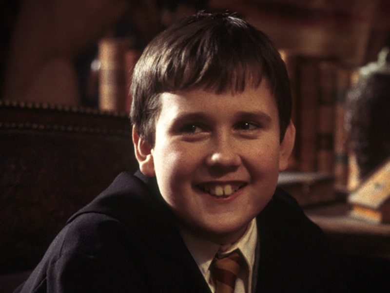 First-year Neville Longbottom smiling and looking off into the distance in his class robes and red and yellow Gryffindor tie in the Hogwarts library.