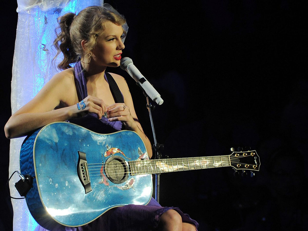 Taylor Swift performing in a purple halter dress with a blue guitar during her "Speak Now" era.