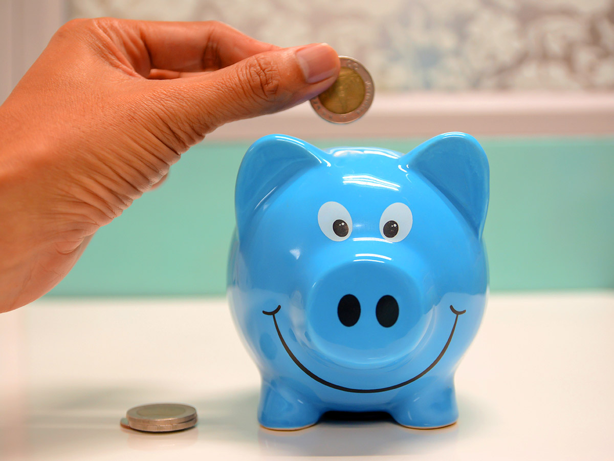 A hand putting a coin into a blue piggy bank with a smiley face.