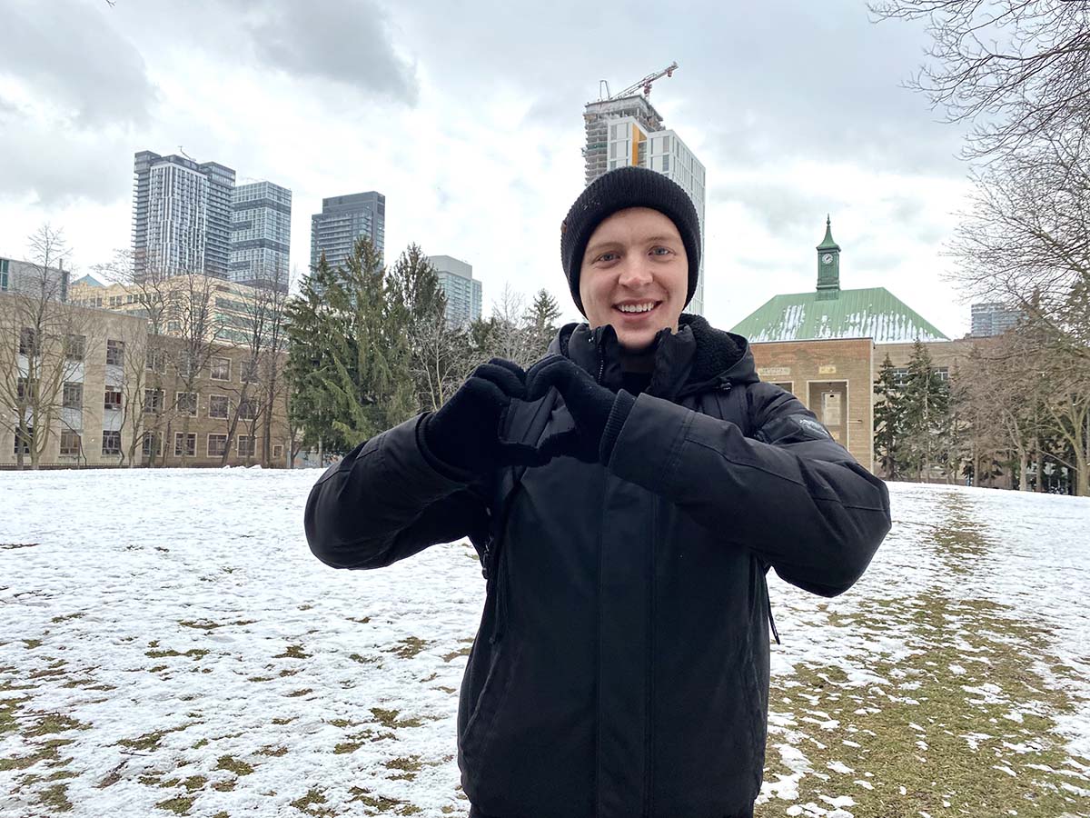 Andrew making a heart with his hands in Kerr Quad with the Toronto skyline in the background.