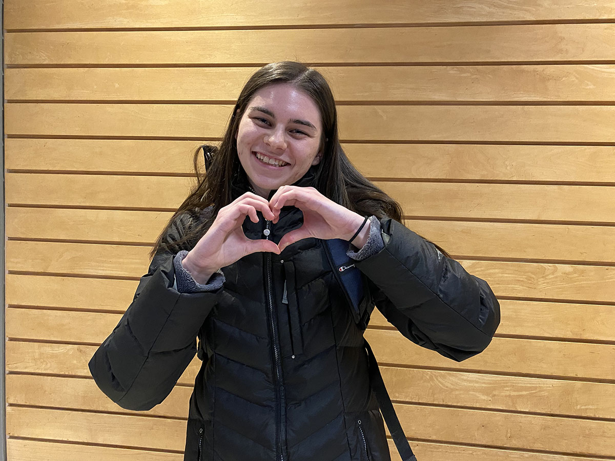 Lauren makes a heart with her hands in front of a wood-panelled wall inside of the George Vari Engineering and Computing Centre.  