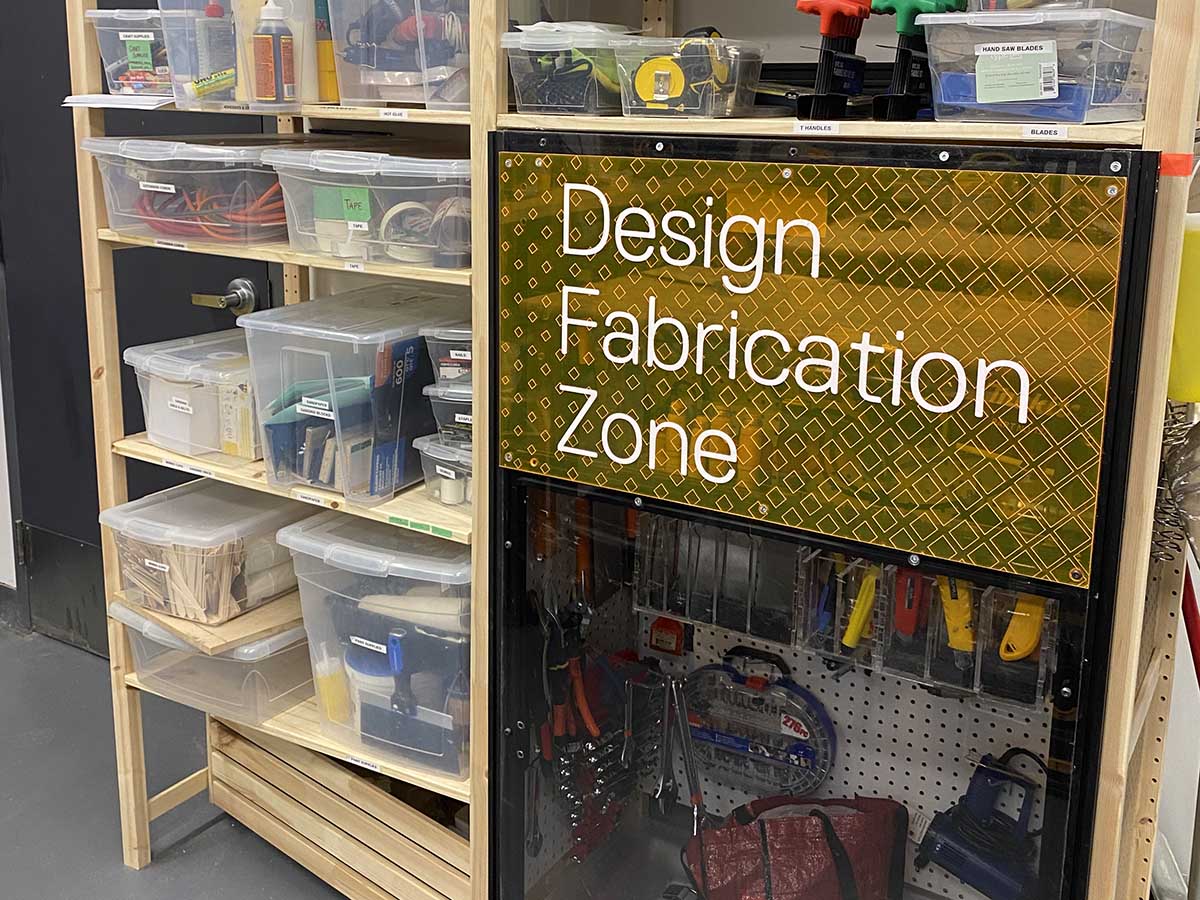 Wooden shelves with see-through containers of supplies and equipment and a sign reading "Design Fabrication Zone."