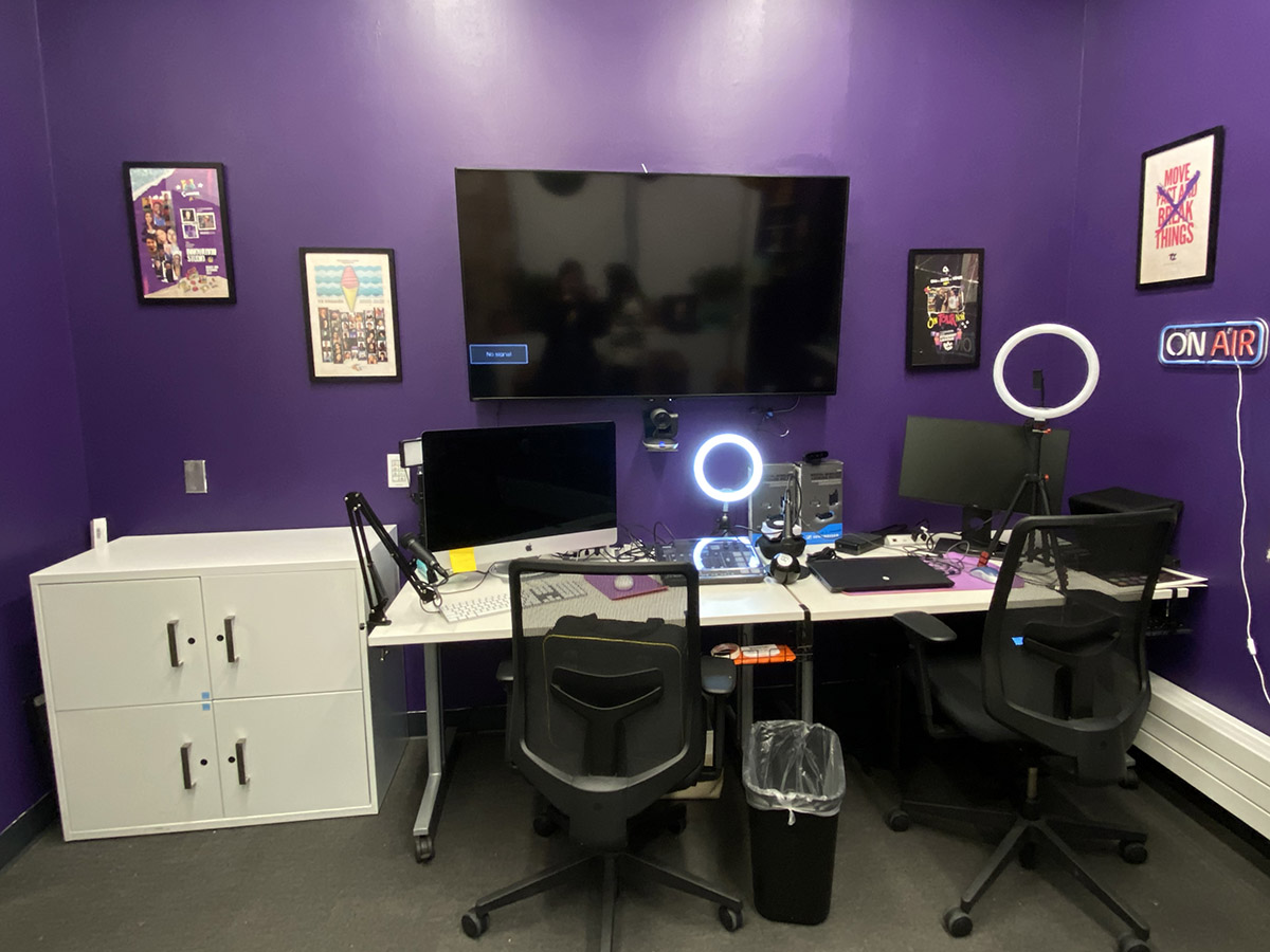 A desk setup with monitors, ring lights and laptops against a purple wall in the Transmedia Zone.