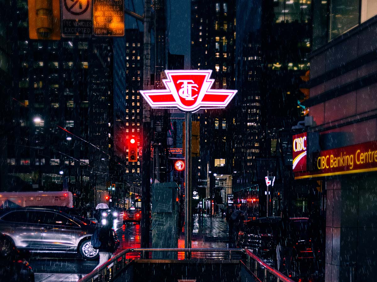 A lit-up TTC sign above a subway stop next to a busy Toronto street at night.