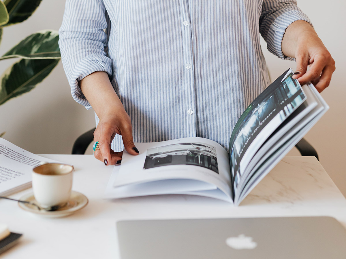 A woman flips through a magazine with a laptop and cup of coffee next to her.