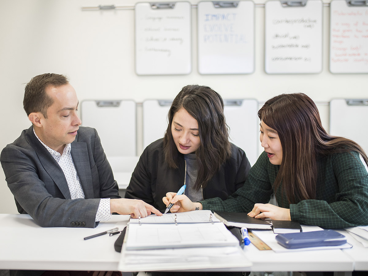 A faculty member sits at a desk and helps two students with their studies.