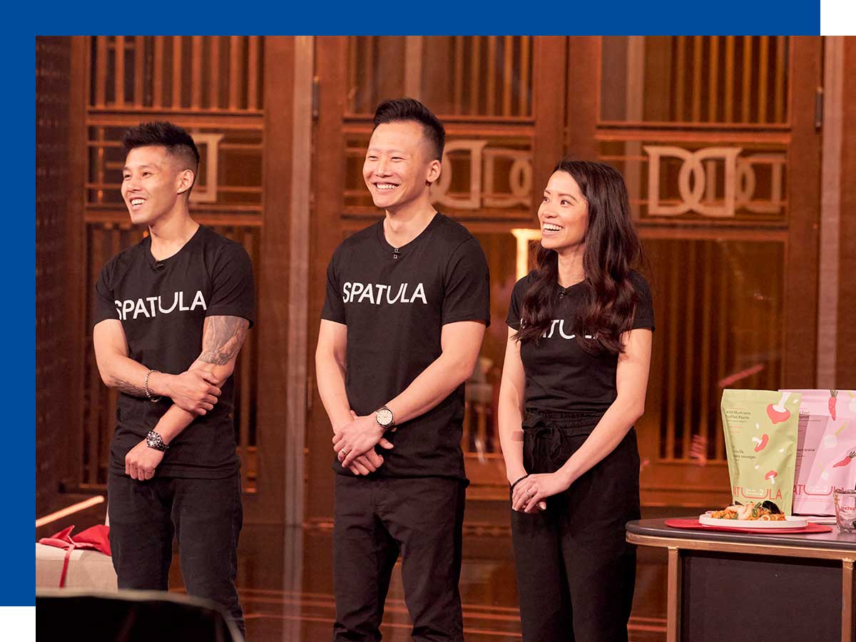 Three Asian Canadians stand before tall wood-toned doors embedded with the Dragons’ Den crest, a cooktop and food display next to them.