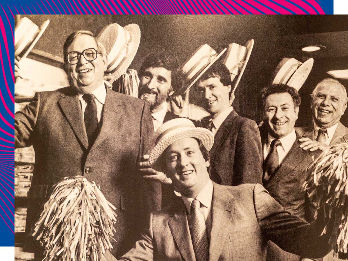 Six staff members of Walter Beauchamp Tailors in 1963 wear suits and raise their hats. One staff member in the centre front holds pompoms.