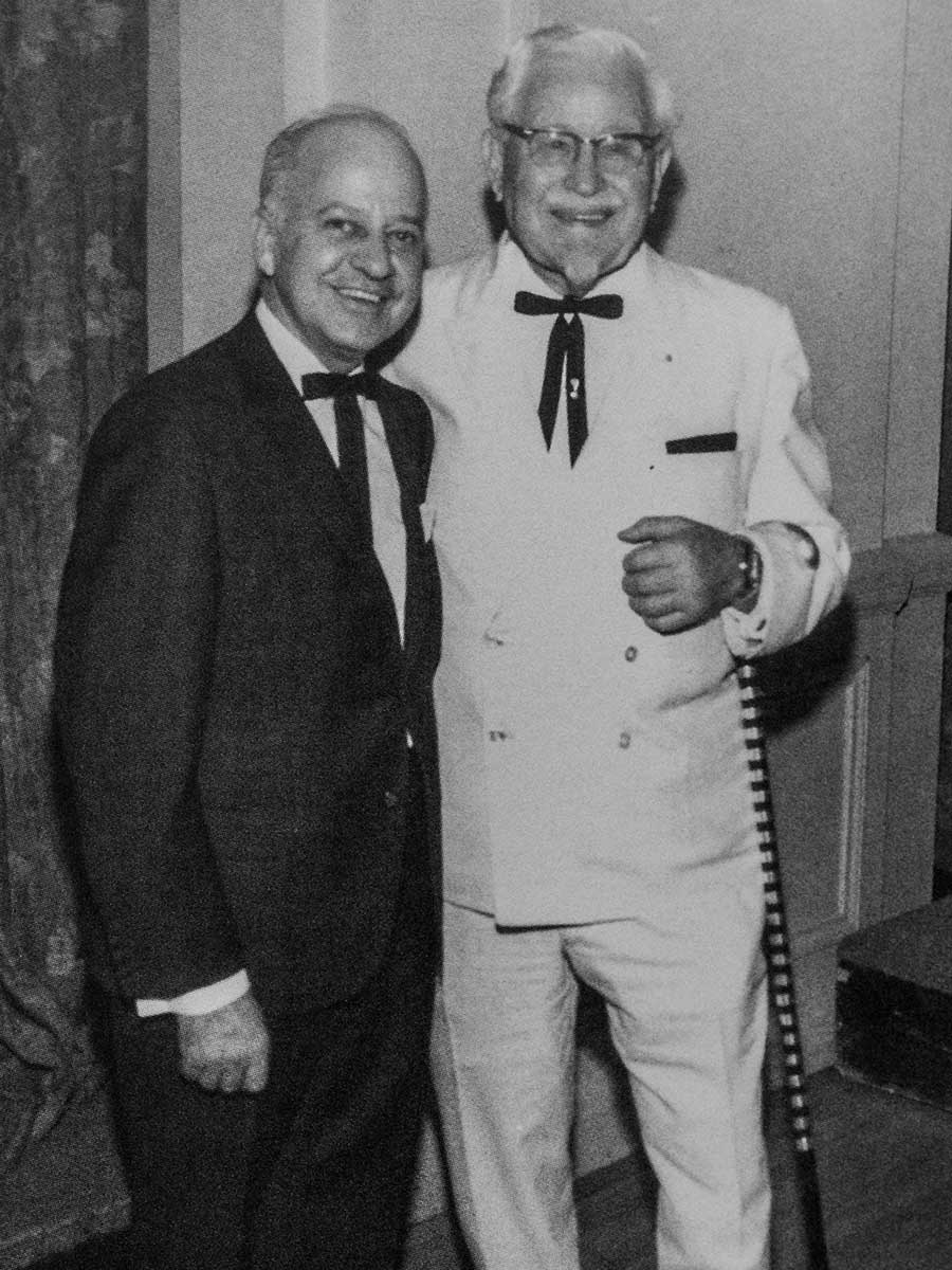 Colonel Harland Sanders of KFC stands wearing a white double breasted suit. Next to him, in contrast,stands Merton Chester wearing a dark suit.