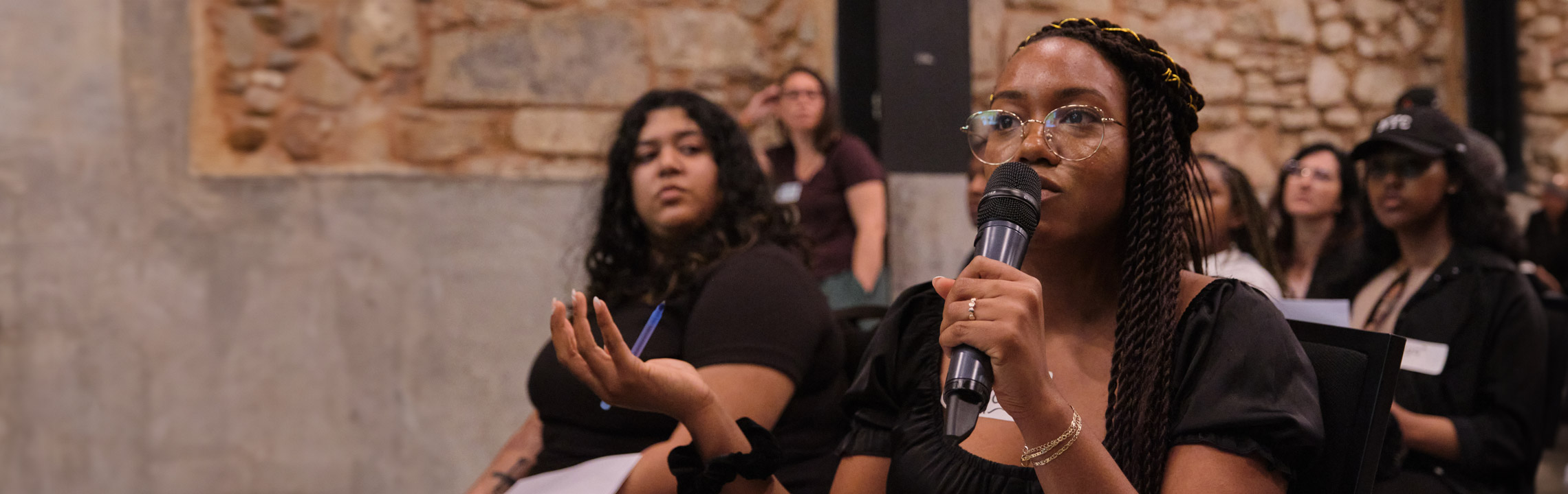 Woman speaking with a mic while seated in an audience 