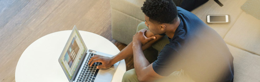 A student works on his laptop with his backpack and phone resting next to him on a couch.