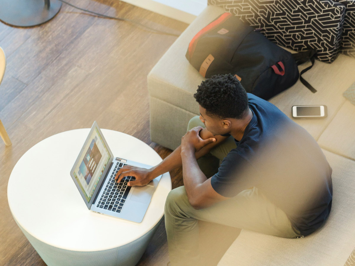 A student works on his laptop with his backpack and phone resting next to him on a couch.