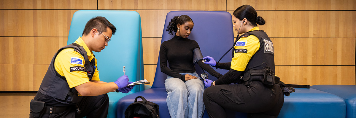 Two TMU Security emergency personnel take vital signs of a student in a DCC lounge area.