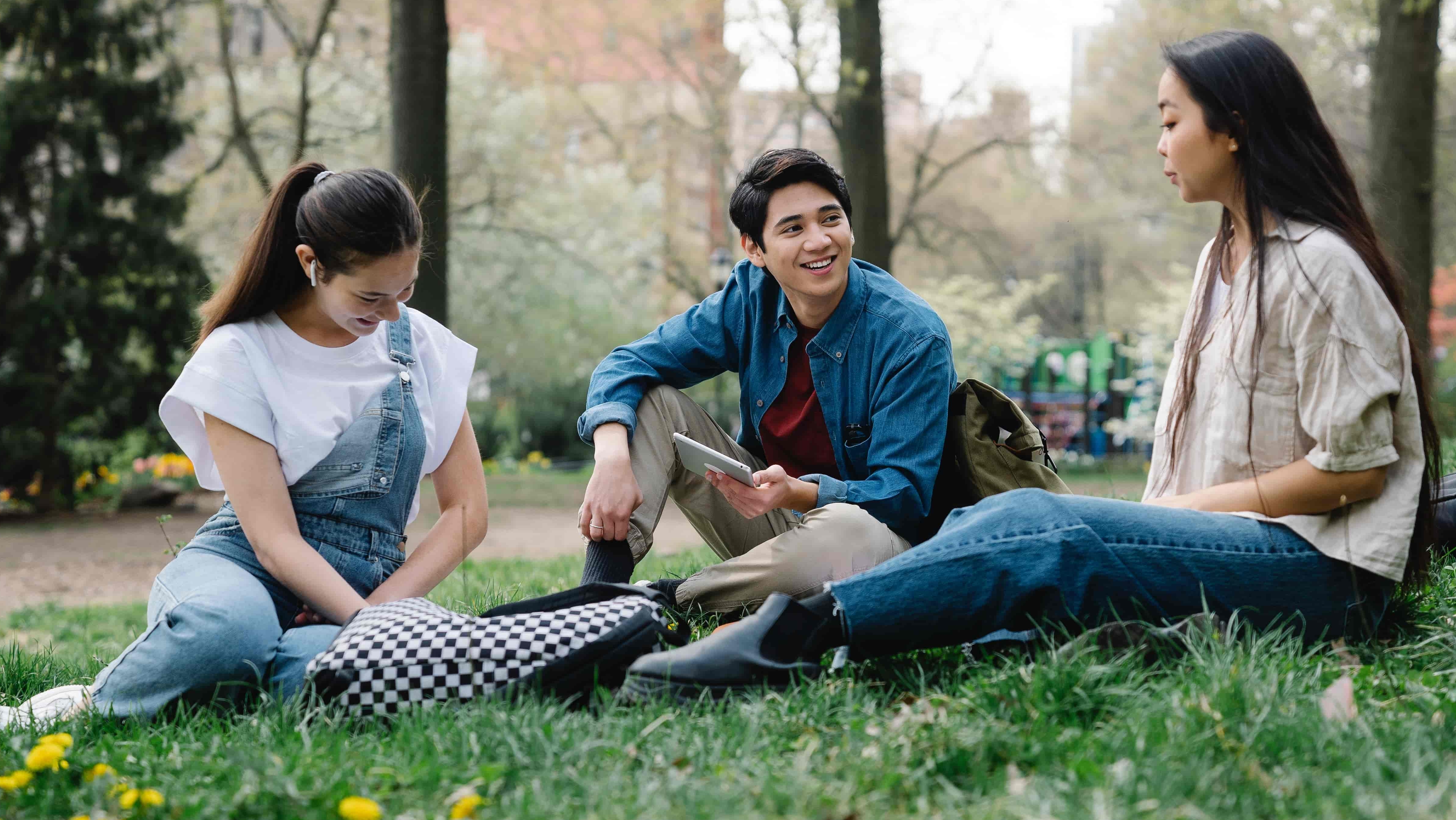 Three students sitting in a semi-circle on the grass having a lighthearted conversation.