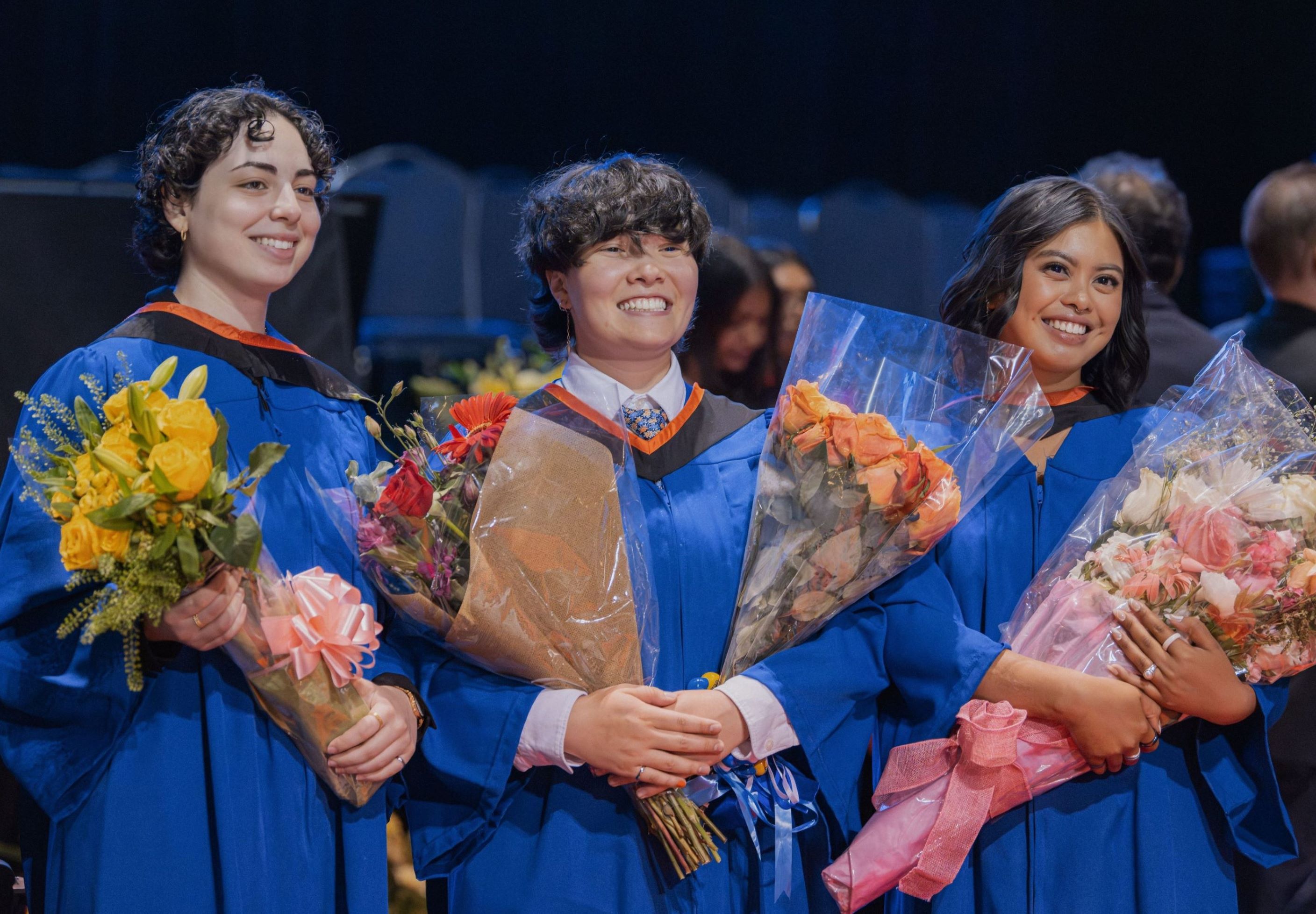 Three graduates smiling and posing for a photo holding bouquets of flowers.