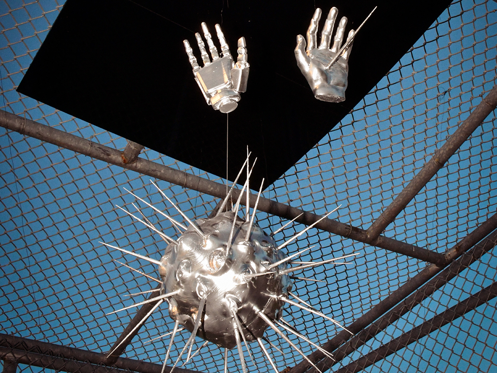 A metallic sphere of eyes and spikes is suspended under metallic hands. There is a chain-link fence in the background.