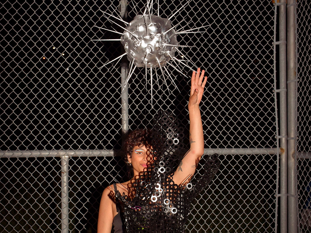 A 3D printed and laser cut sculpture of a portion of a female body is suspended under a metallic spikey sphere and in front of someone with their hand raised in front of a chain link fence.