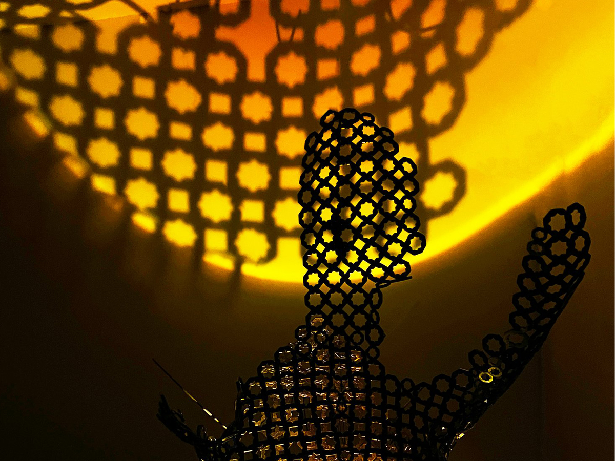 A zoomed-in portion of an intricately-cut mesh sculpture over a bright orange backdrop. The shadow