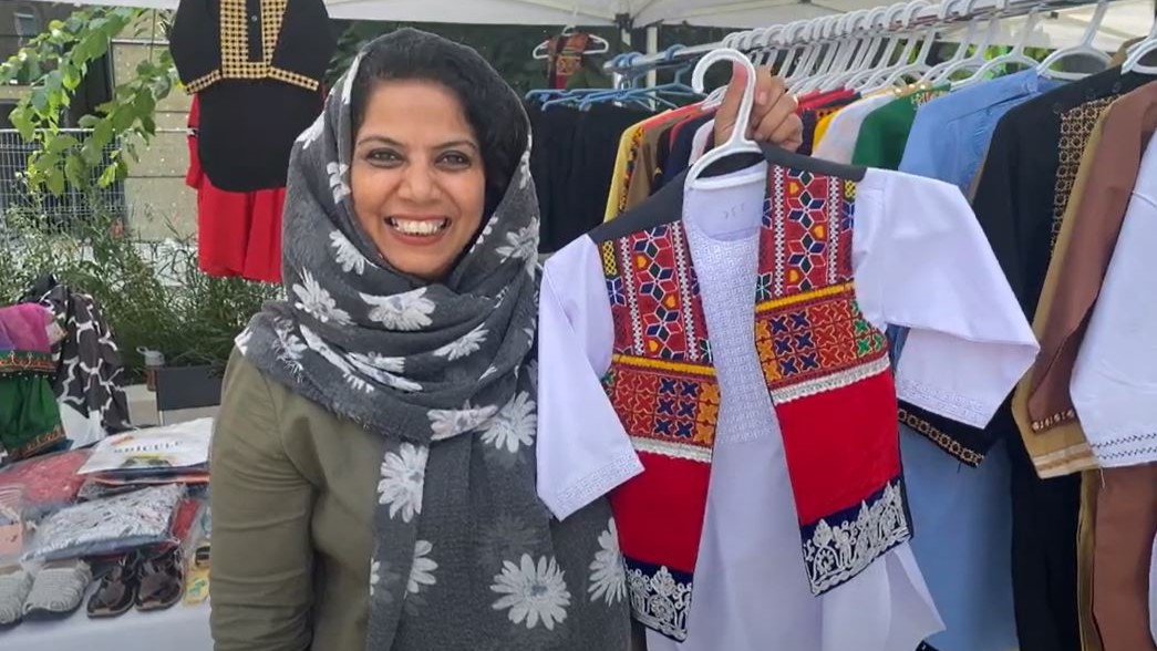 A racialized woman wearing a grey head scarf holds up a boy’s outfit, a red vest with embroidery detailing layered on top of a long-sleeve white shirt.