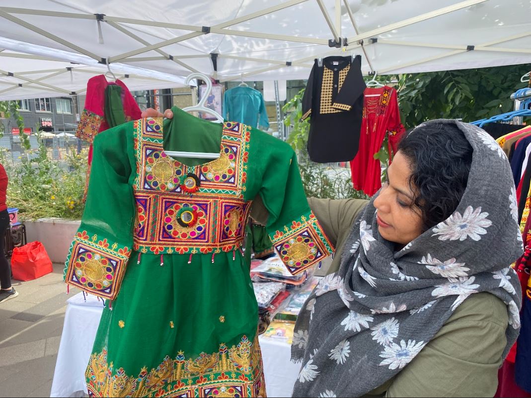 A green dress with highly detailed colourful embroidery is displayed by a racialized woman entrepreneur wearing a grey head scarf.