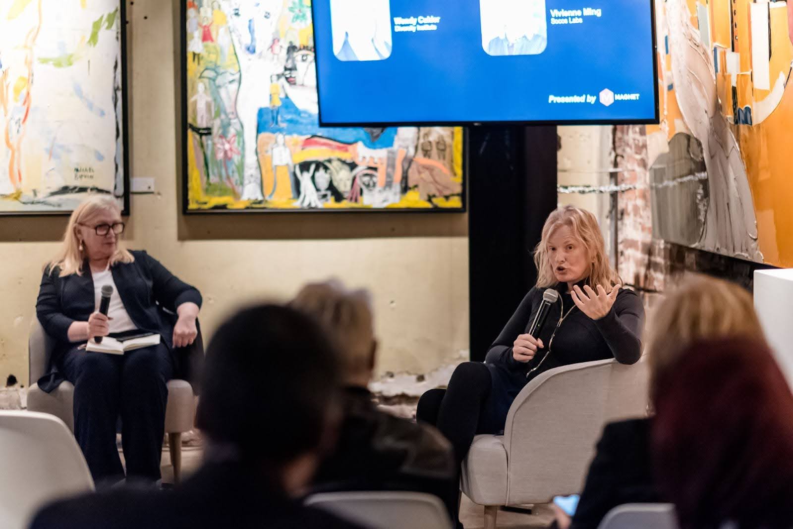 Two women, one speaking into a handheld microphone, sitting on chairs on stage during an event at an art gallery venue. 