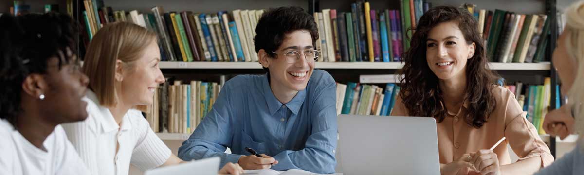 Five TMU community members sit around a table in the library as they discuss the EDI strategy and action plan. One student, dressed in a long-sleeved peach shirt with a laptop in front of her, smiles at another student across the table. Another student wearing glasses and a long-sleeved blue shirt holds a pen, smiling, as he listens to input from his peers.