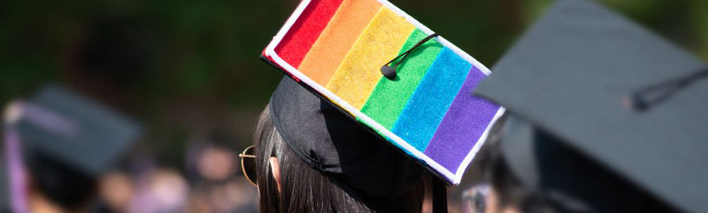 A graduate wearing a square academic cap in the colours of the Pride flag, red, orange, yellow, green, blue and purple.