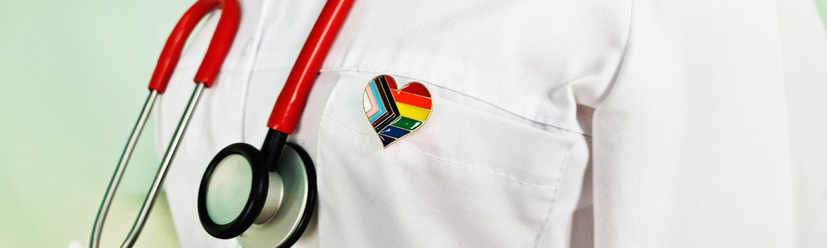 Close-up of a medical professional wearing a white coat and stethoscope, featuring an enamel pin in the shape of a heart with the Progress Pride flag.