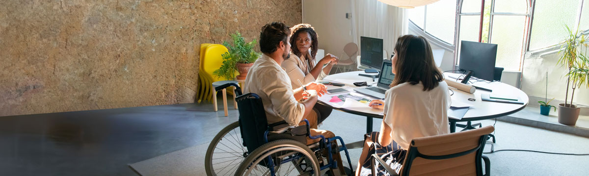 Three people talking and working at a table. Two people are in office chairs, and one is in a wheelchair.