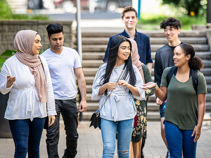 Five diverse students walking outside. Two students in the foreground are talking.