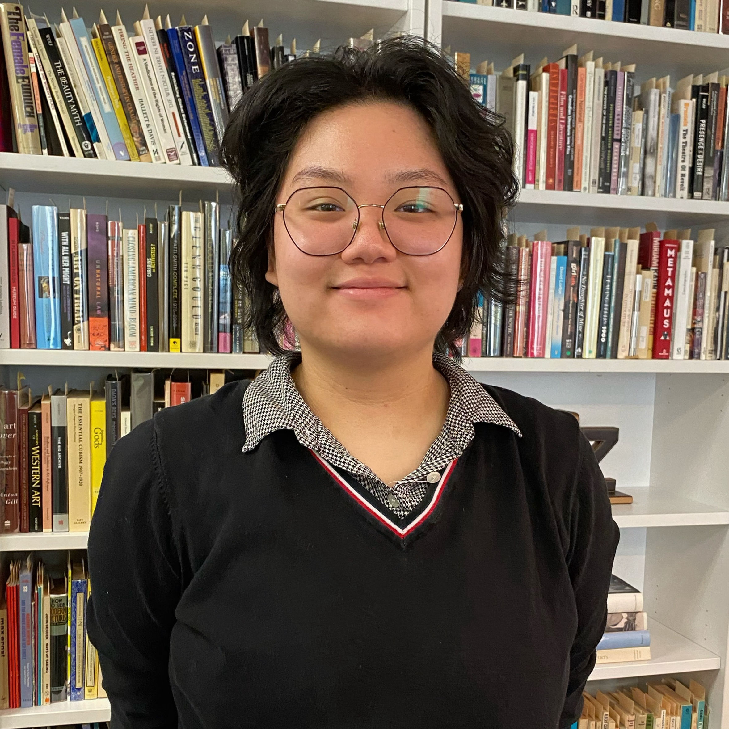 Headshot of Student Staff, Max Huynh smiling with their hands behind their back in front of a bookshelf