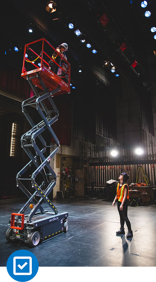 Two students on stage in the Ryerson Theatre. One student is operating a scissor lift while the other is providing direction. Both are wearing hard hats, vests and boots. 