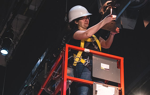 A student working on a scissor lift to adjust lights on the Ryerson stage. They are wearing a harness and hard hat.