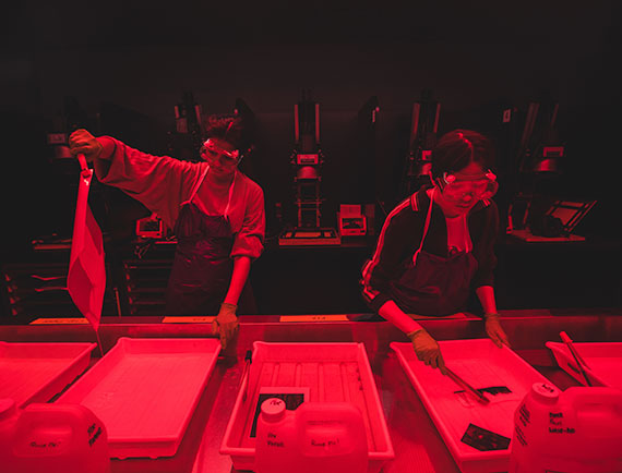 Two students under red lights developing photos in a darkroom on campus.