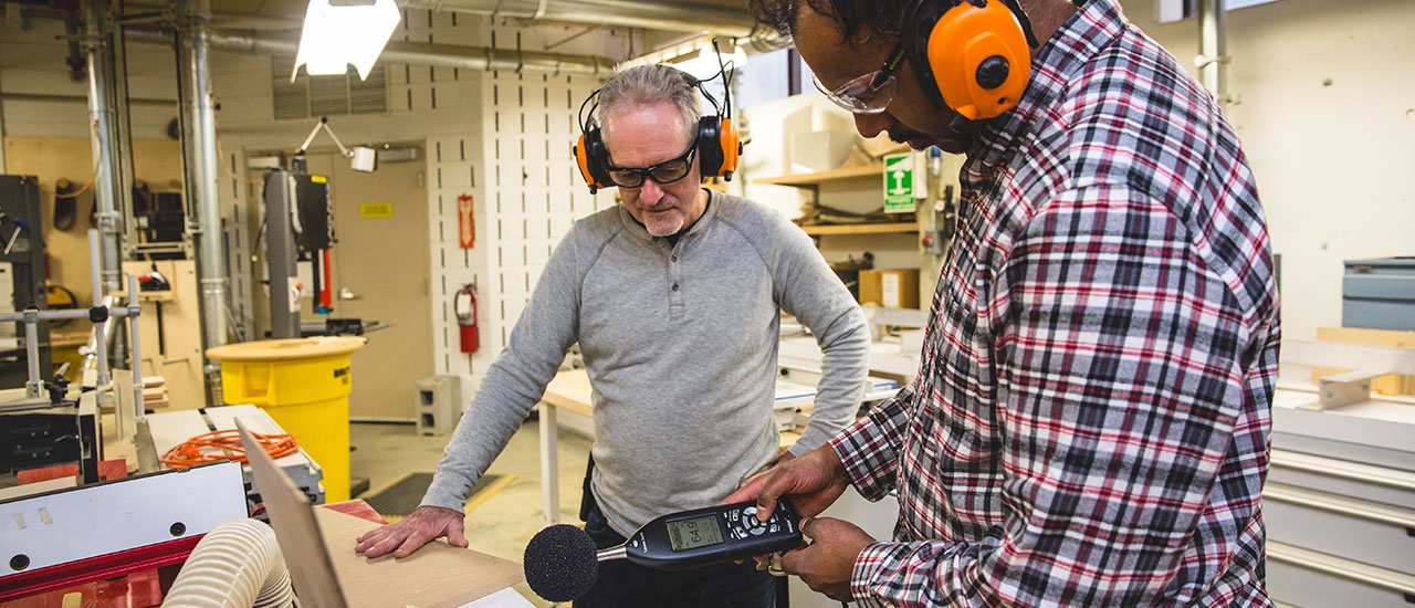 A member of the Environmental Health and Safety team in a workshop reviewing a noise metre with a departmental safety officer. Both are wearing over-ear hearing protection. 