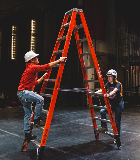 Two students about to use a ladder. One is climbing the first step while the other is providing support, both are wearing hard hats.