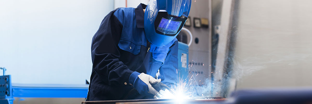 A person wearing a welder's face shield, gloves and protective coveralls uses a torch to cut a band of metal.