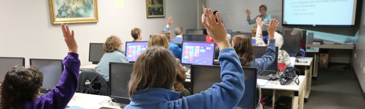 An instructor stands at the front of a room, participants sit behind computers, some have their hands up.