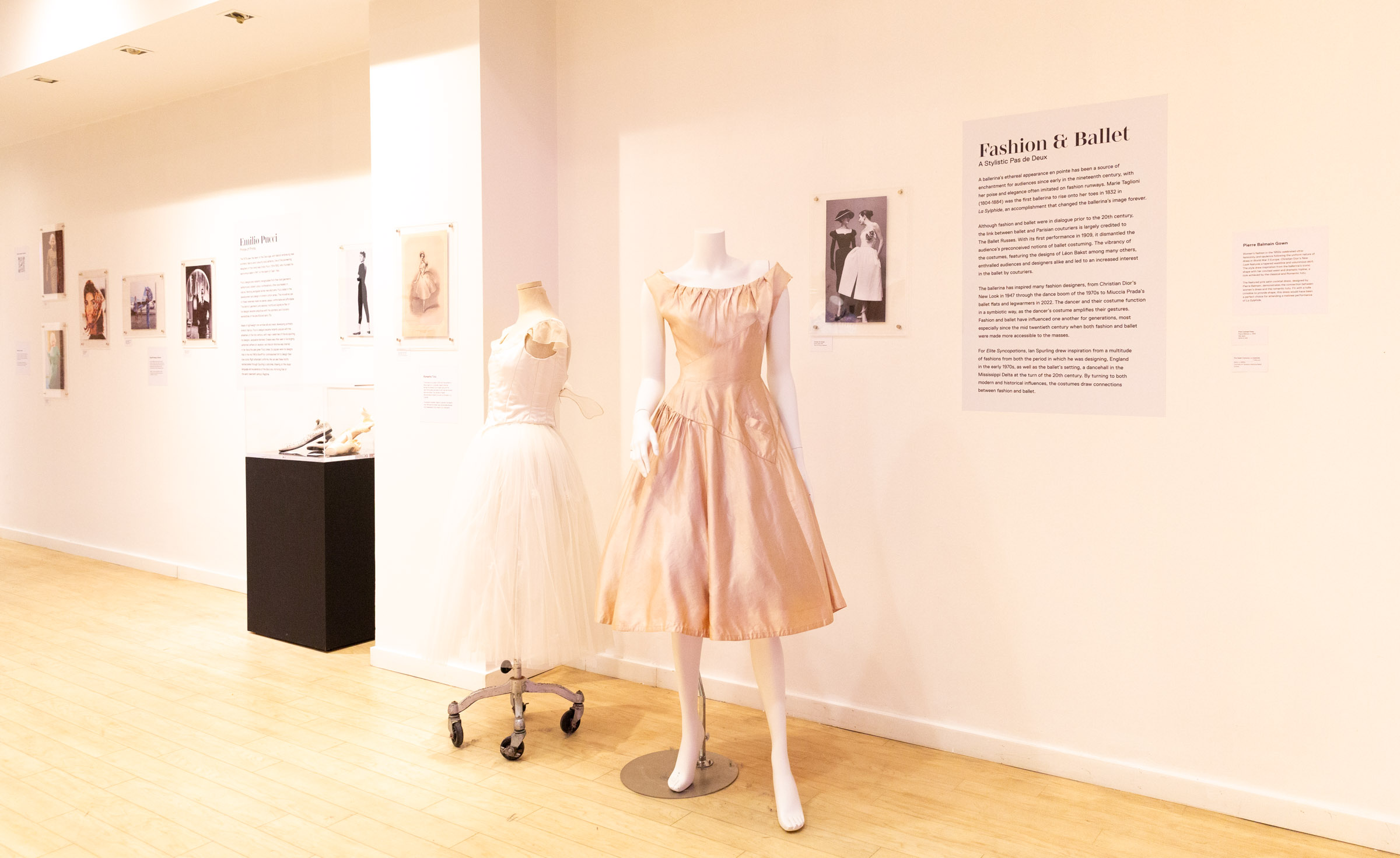 Two dresses, a white and pink are displayed next to text on the wall that reads "Fashion & Ballet". 