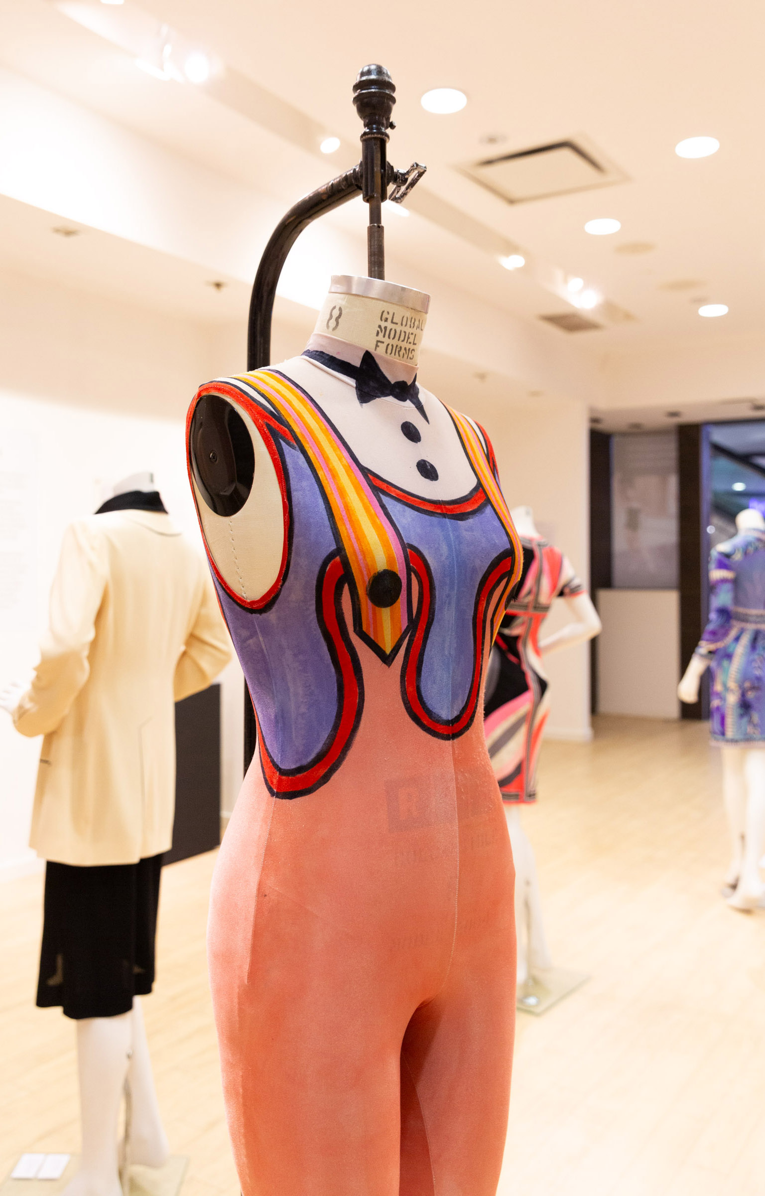 A close up of a trompe l'oeil costume in pink, orange, blue, black and white depicting pants with suspenders over a shirt with a necktie. 