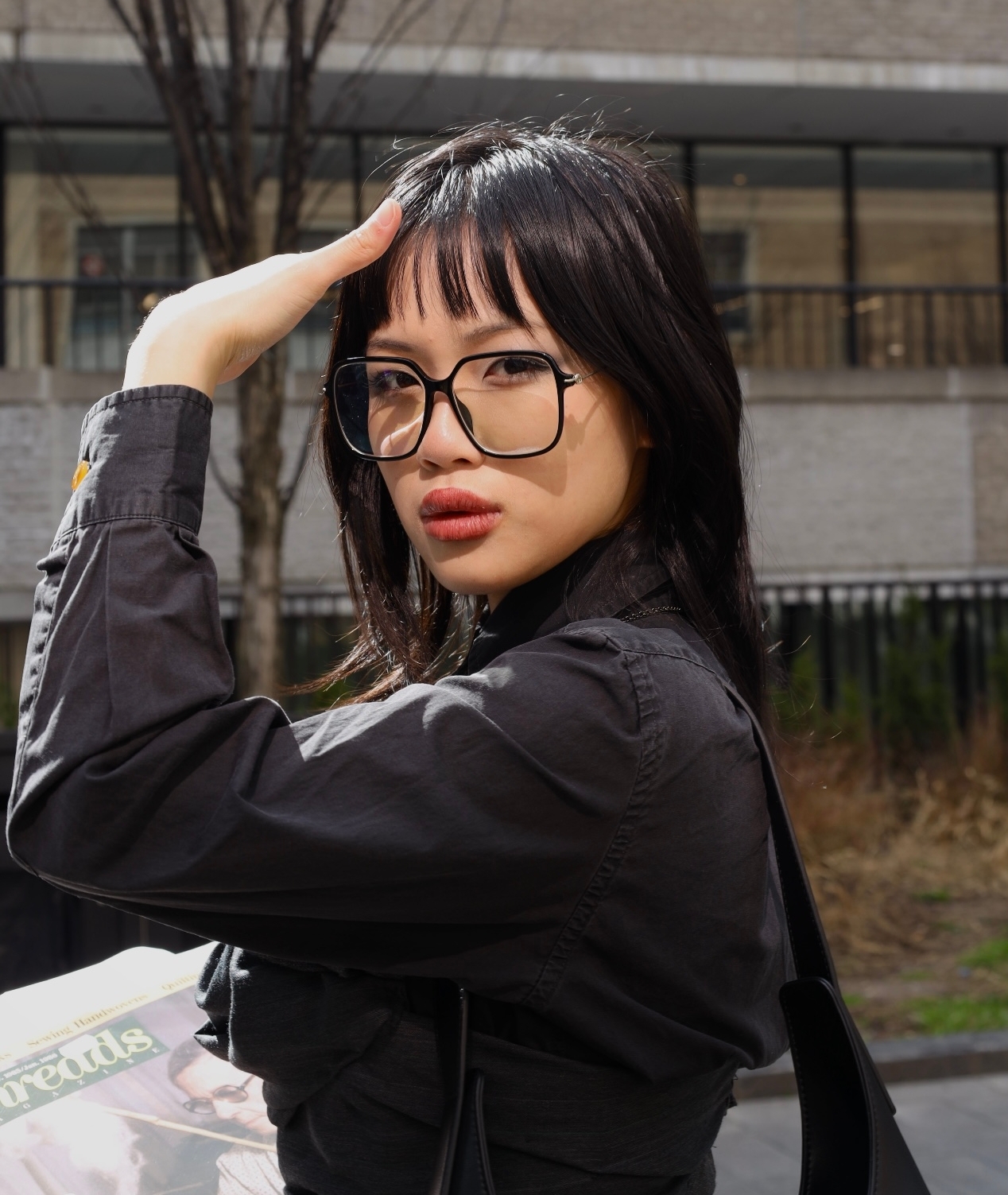 Headshot of Elysia outside. Elysia is an East Asian woman with black hair wearing large glasses. Her hand is shielding her face from the sun as she looks at the camera. 