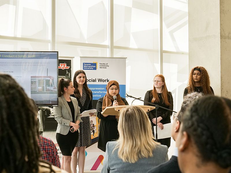 Five students stand behind a podium addressing an audience.  Farah Fatima holds the microphone