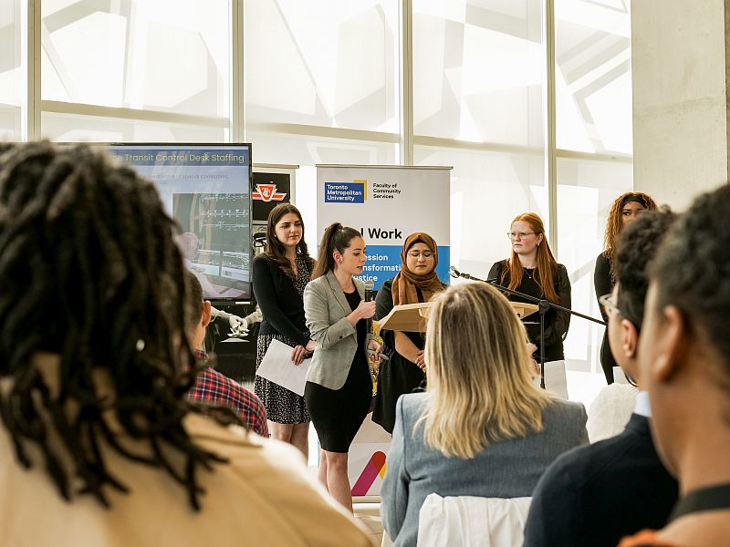 Five students stand behind a podium addressing an audience.  Madalina Beloia-Cheres holds the microphone