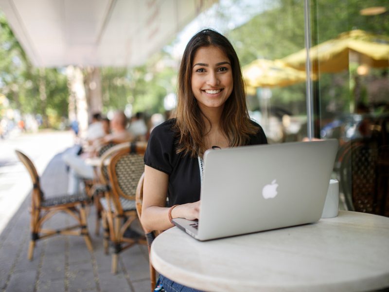 Female graduate student sitting on Balzac's patio typing on laptop and smiling