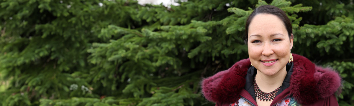 Muckpaloo Ipeelie smiles in front of green evergreen trees, she is wearing a maroon jacket with a fur collar and a beaded necklace.